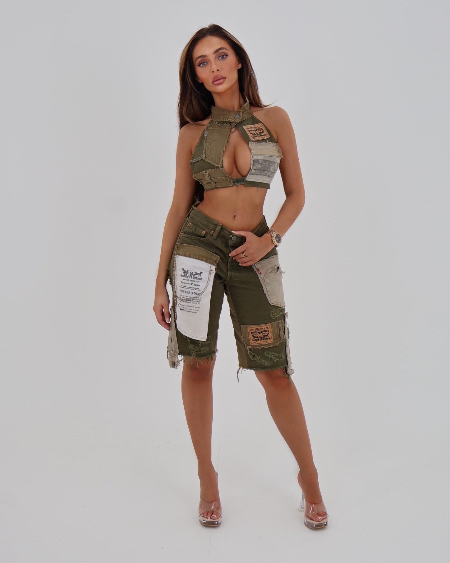 A woman modeling a matching military-inspired two-piece set consisting of a halter crop top and cargo shorts, both in olive green with beige patches and white printed labels, against a light gray studio background.