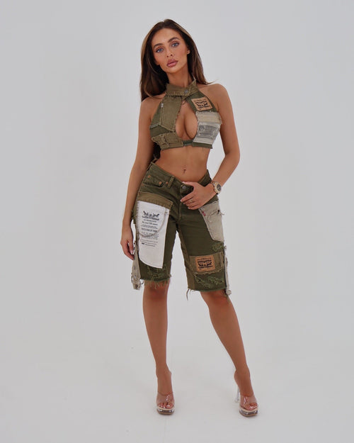 A woman modeling a matching military-inspired two-piece set consisting of a halter crop top and cargo shorts, both in olive green with beige patches and white printed labels, against a light gray studio background.