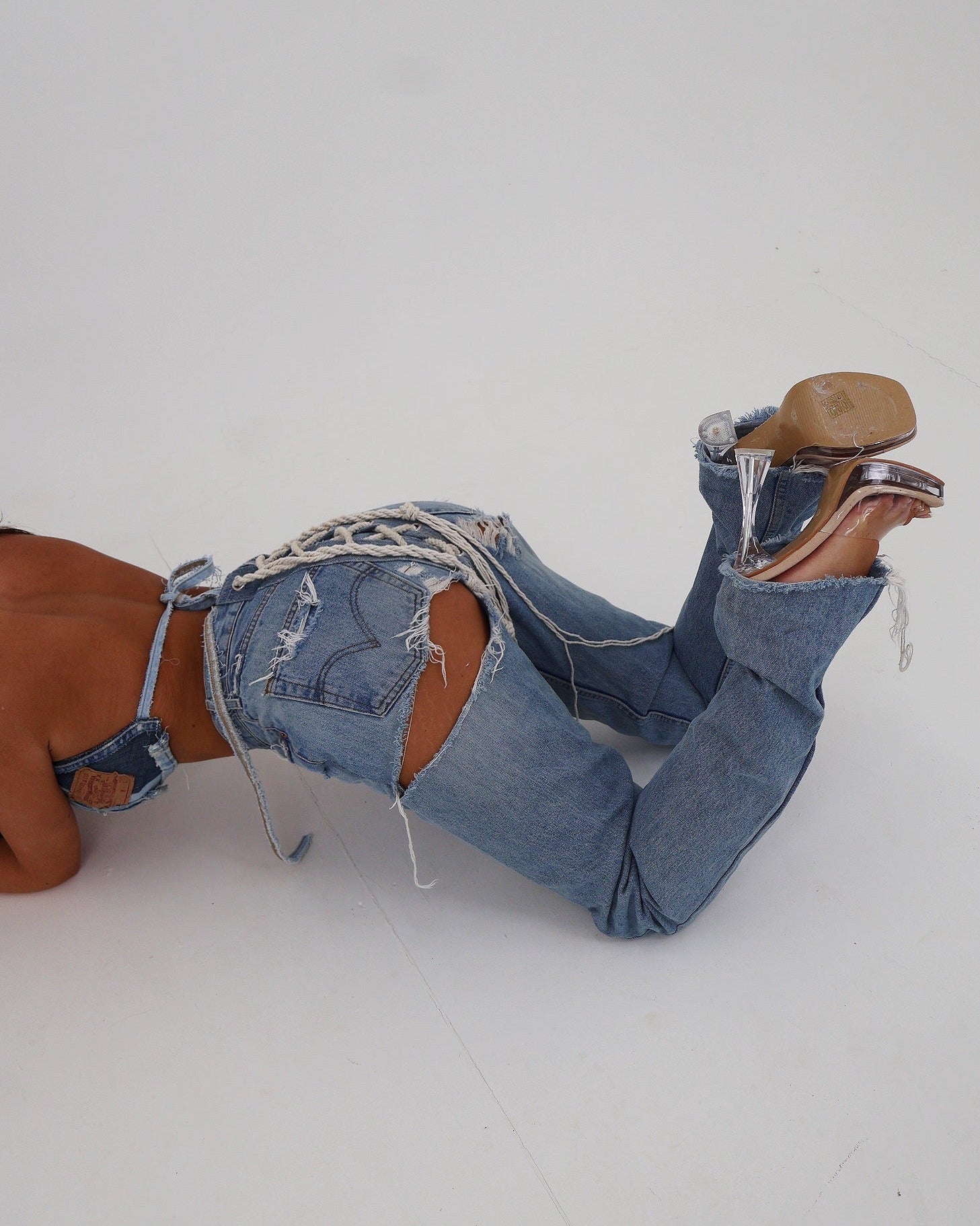 A person lying on a white studio floor wearing distressed light blue denim jeans with ripped details, a matching blue denim headband, and brown heeled sandals.